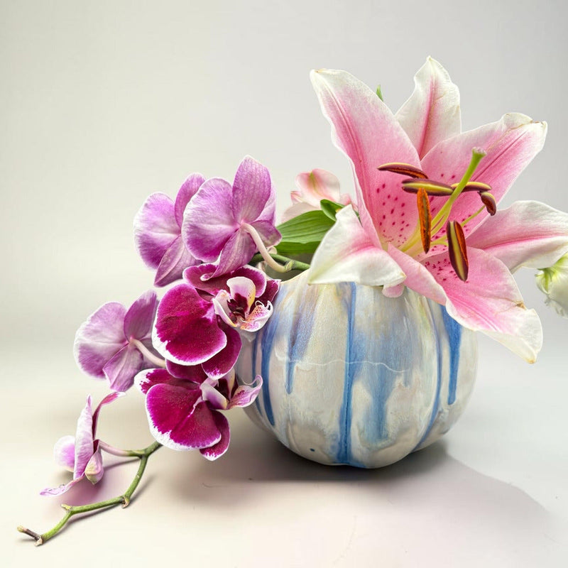 A decorative ceramic vase with blue and white vertical stripes, filled with pink and white lilies and purple orchids, set against a clean light gray background.