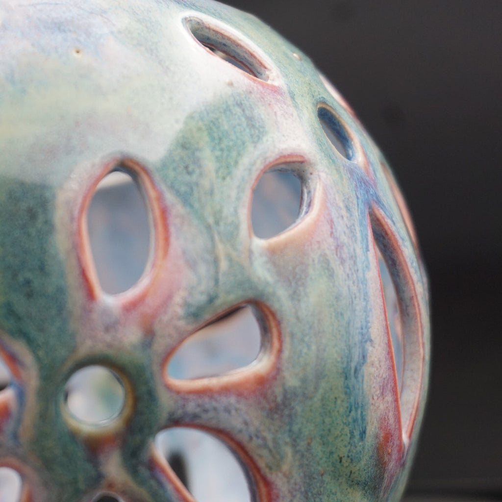 Close-up of a decorative ceramic vase with a perforated, lotus-like pattern. The glossy glaze features a marbled blend of teal, green, blue, and terracotta orange-red tones against a dark, out-of-focus background.