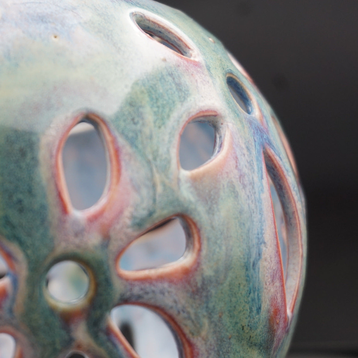 Close-up of a decorative ceramic vase with a perforated, lotus-like pattern. The glossy glaze features a marbled blend of teal, green, blue, and terracotta orange-red tones against a dark, out-of-focus background.