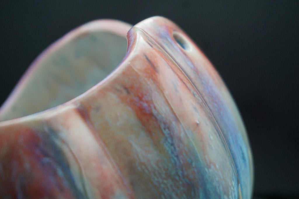 Close-up of a ceramic bowl with a marbled, iridescent finish showing shades of pink, blue, purple, and beige against a dark background.