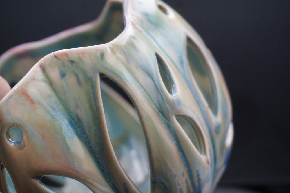 A close-up of a sculptural ceramic vase with organic, organic-shaped cutouts and a glossy marbled glaze in soft beige, blue, and green tones against a dark background.