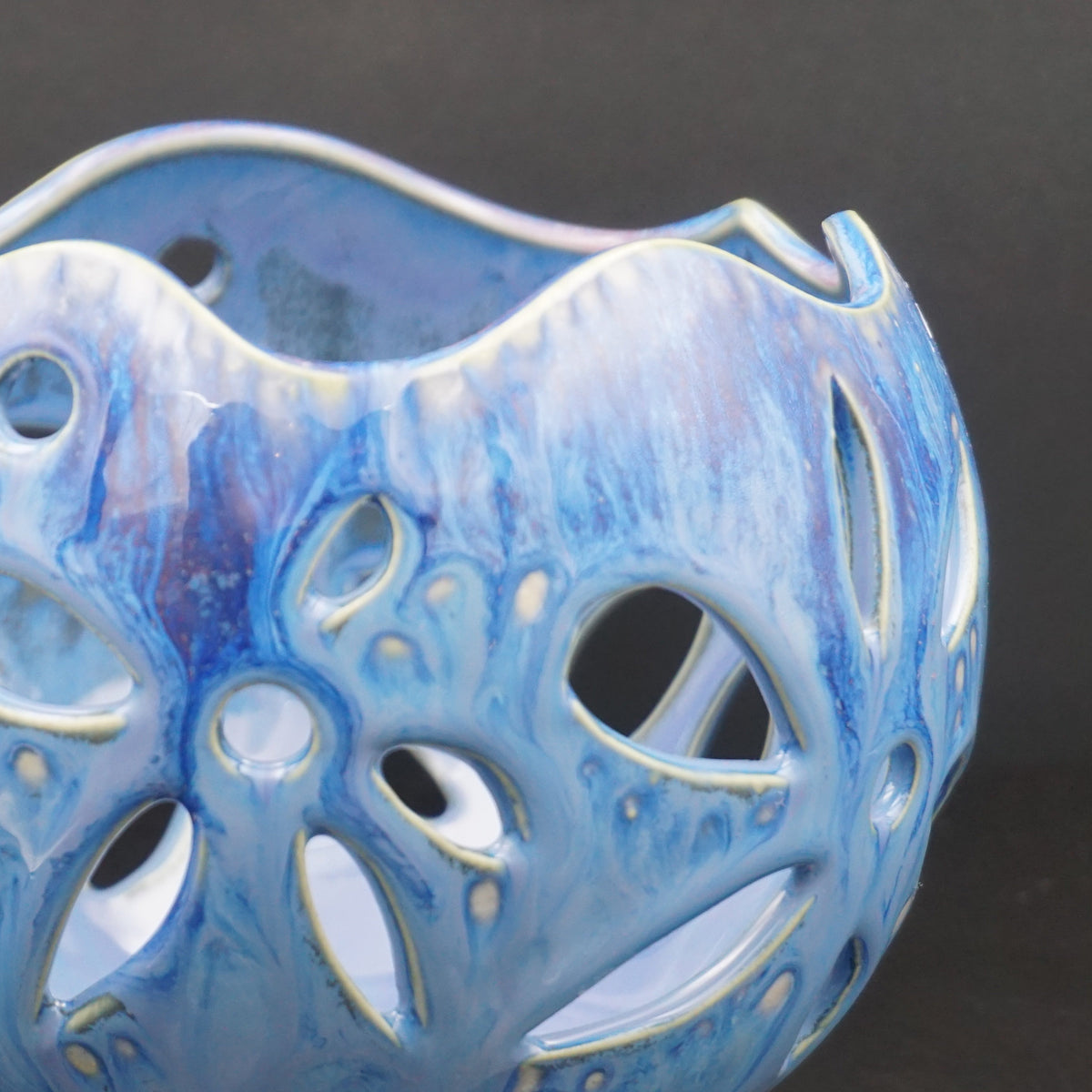 A close-up of a decorative ceramic vase with a blue and white marbled glaze and an intricate openwork pattern, set against a dark gray background.