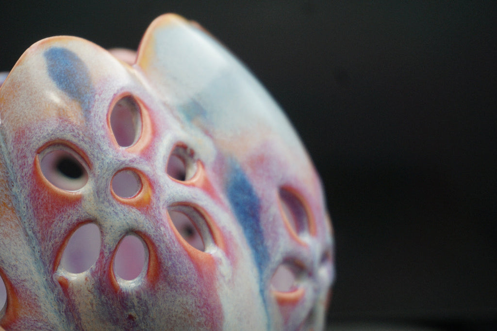 A close-up of a decorative ceramic object with a porous, lotus-like pattern, featuring a marbled glaze of pink, blue, orange, and cream against a dark background.