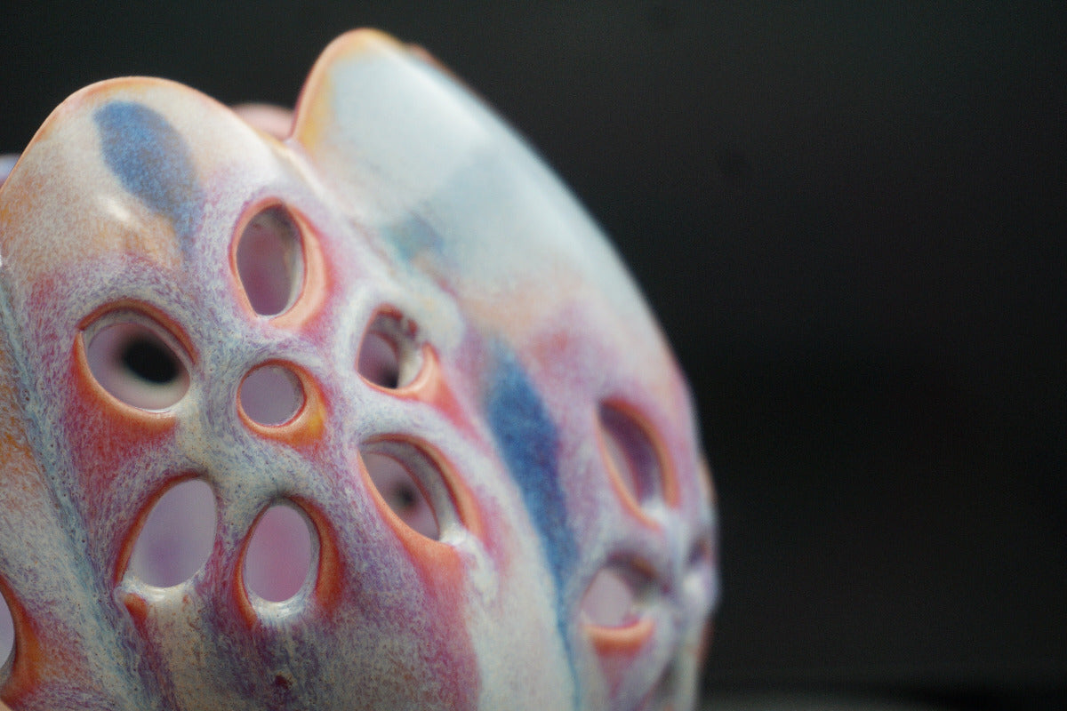 A close-up of a decorative ceramic object with a porous, lotus-like pattern, featuring a marbled glaze of pink, blue, orange, and cream against a dark background.