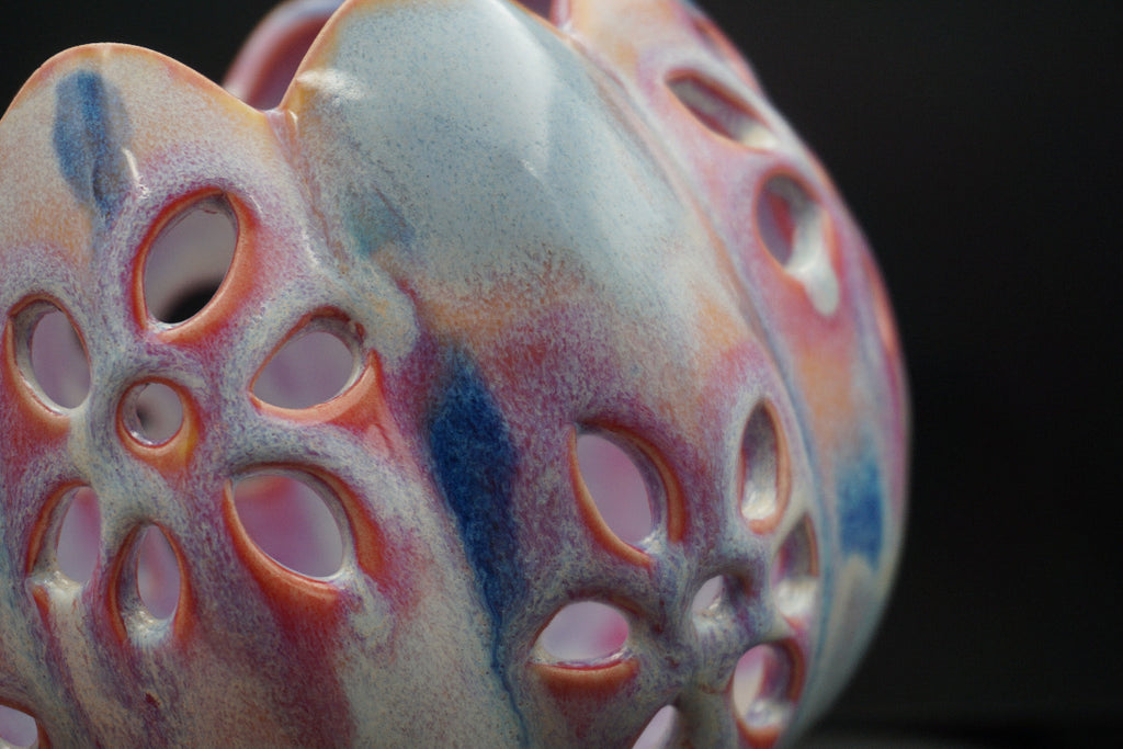 A close-up of a ceramic vase with a textured, porous surface featuring a marbled glaze of pink, blue, orange, and white on a black background.