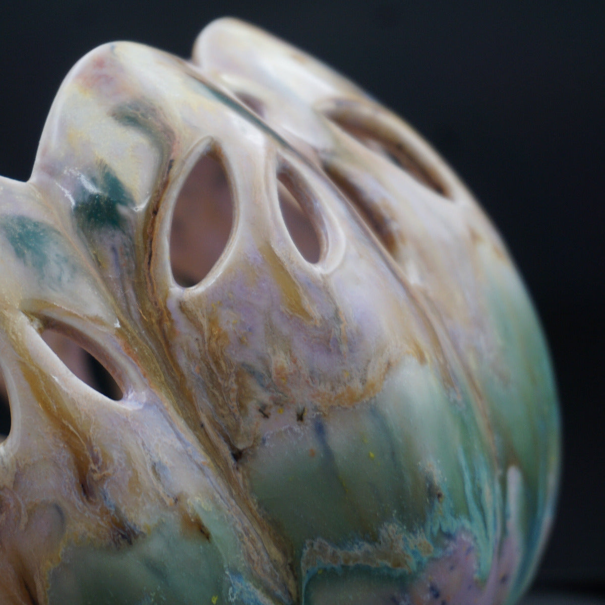 A close-up of a sculptural ceramic vessel with organic, shell-like curves and multiple eye-shaped cutouts, featuring marbled glazes in cream, tan, green, and soft pink against a dark background.