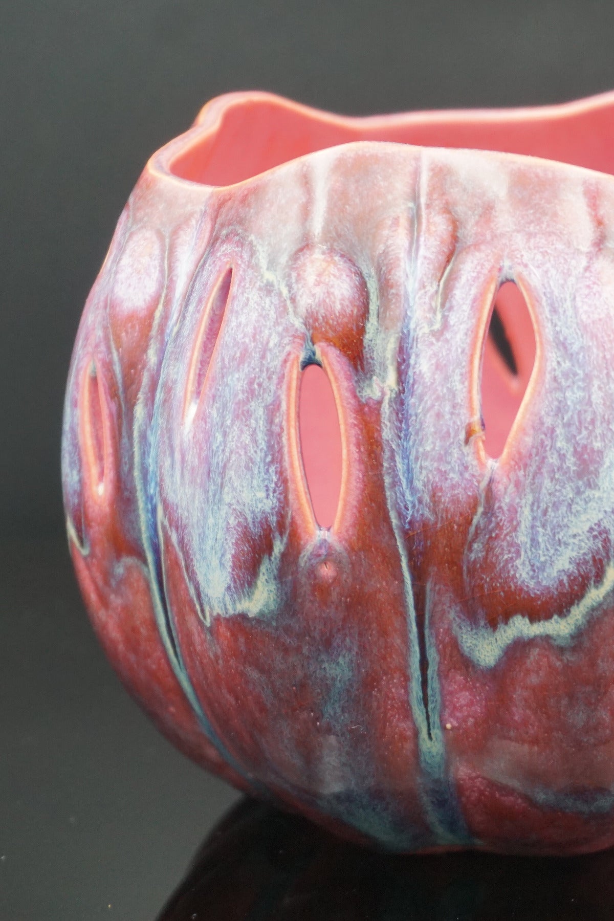 A close-up of a ceramic bowl with a glossy, marbled glaze in pink, red, and blue tones, featuring irregular cutouts along the side.