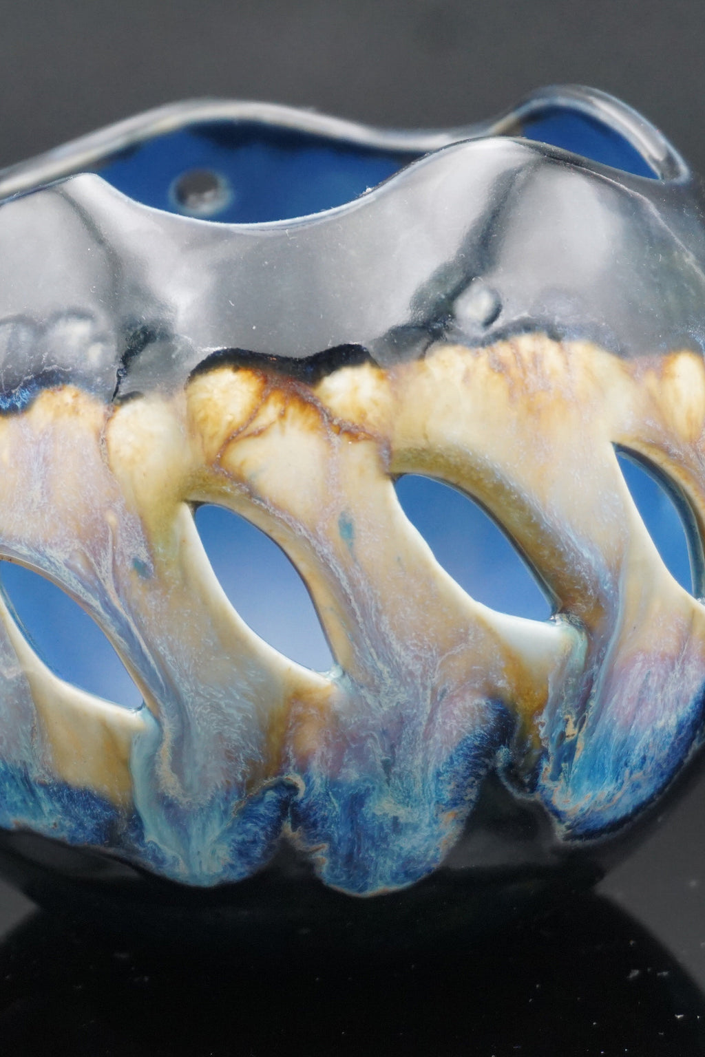 A close-up of a sculptural glass bowl with organic, wavy cutouts, showcasing a glossy finish with blue, cream, and brown marbled patterns.