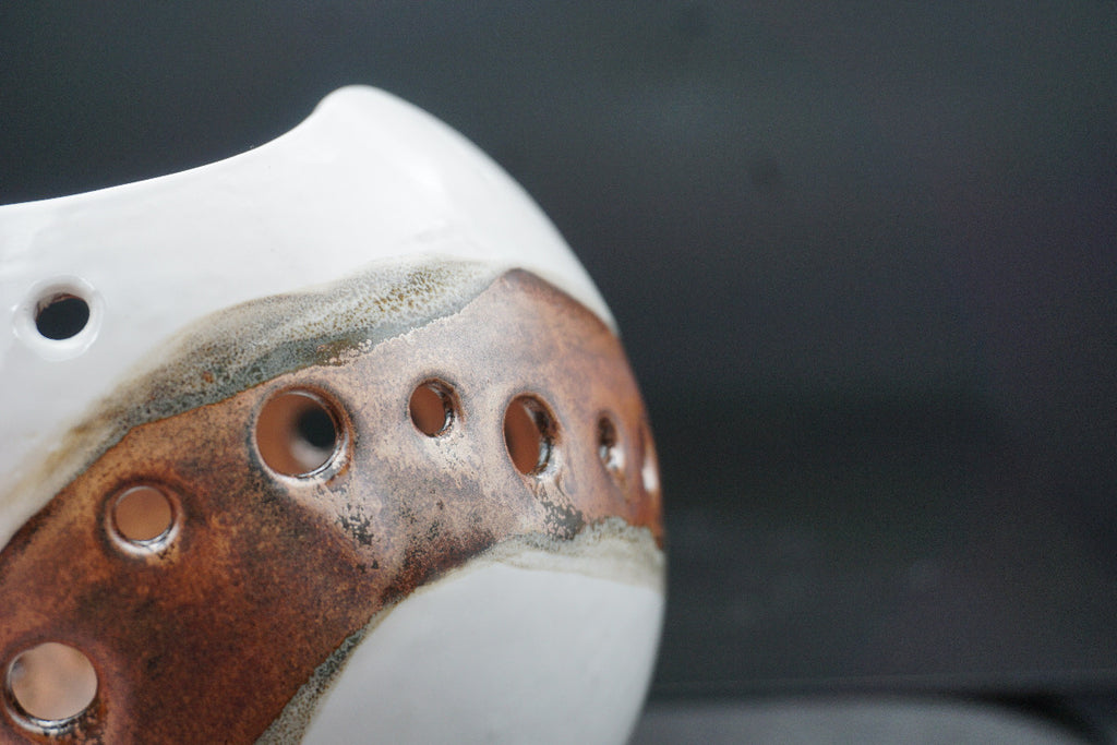 Close-up of a ceramic vessel with a glossy white exterior and a rustic brown glazed section featuring multiple circular cutouts, set against a dark gray background.