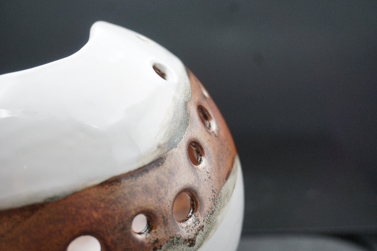 Close-up of a ceramic vessel with a glossy white upper section and a rustic brown lower section featuring circular cutouts, set against a dark gray background.