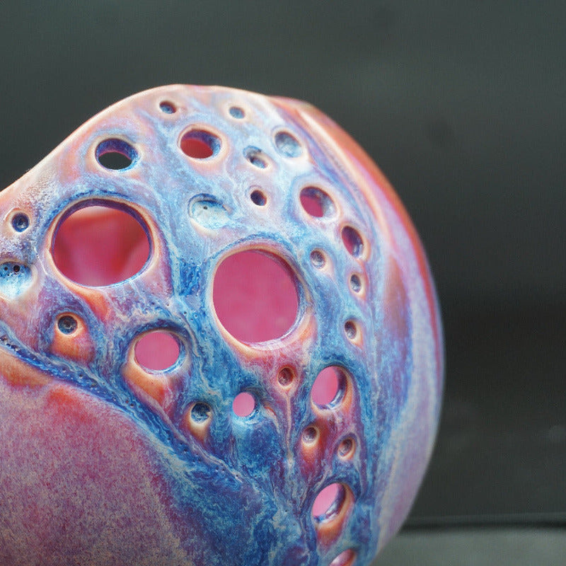A close-up of a hand holding a sculpted ceramic vessel with a glossy finish, featuring organic bubble-like cutouts and swirls of pink, blue, orange, and purple on a dark background.