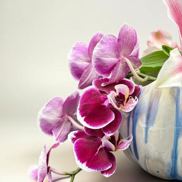 A decorative ceramic vase with blue and white vertical stripes, filled with pink and white lilies and purple orchids, set against a clean light gray background.